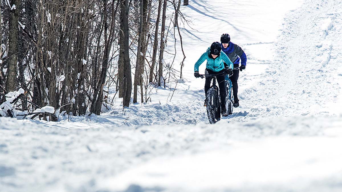 A Female And Male Fatbiker Ride Through A Snow Field