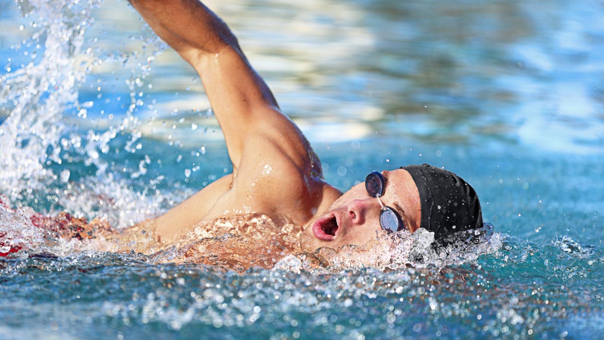 Man Swimmer Swimming Crawl In Blue Ocean Open Water. Portrait Of An Athletic Young Male Triathlete Swimming Crawl Wearing Cap And Swimming Goggles. Triathlete Training For Triathlon.