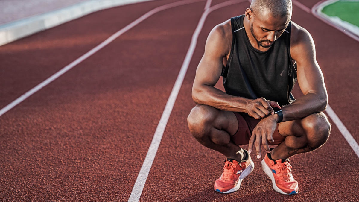 Young Sportsman Sitting On Running Track And Looking At Smartwatch