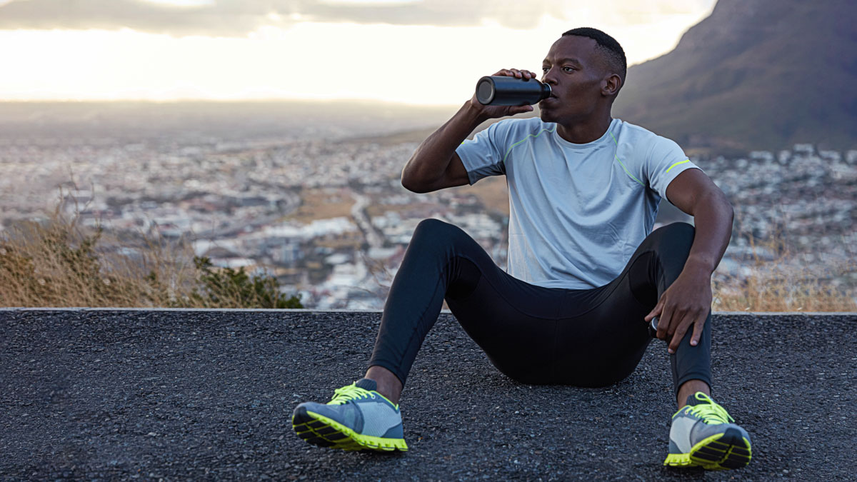 African American Man Drinks Fresh Water From Bottle, Rests On Asphalt, Sits Against Mountain Background Outdoor, Feels Relaxed, Dressed In Casual T Shirt, Sneakes And Trousers. Relaxation Concept