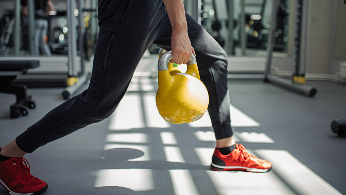 Young Athlete In Black Pants And Orange Sneakers Lifts Yellow Kettle Ball In Gym