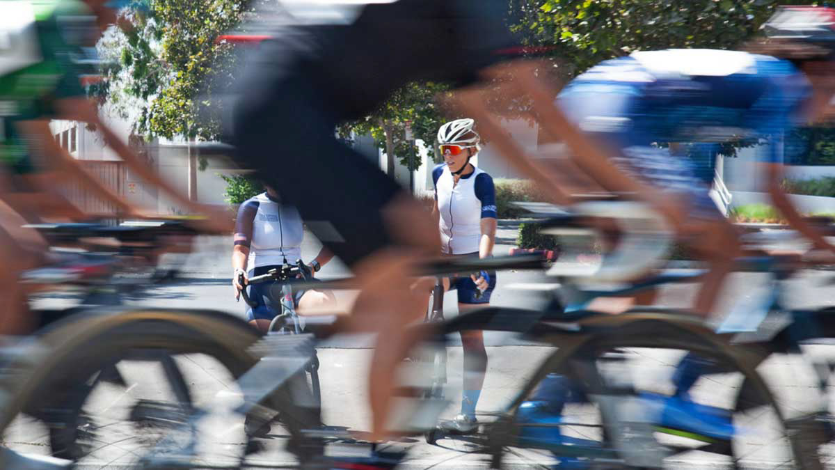 Two Female Cyclists Watch A Crit Race As Riders Blur Through The Frame