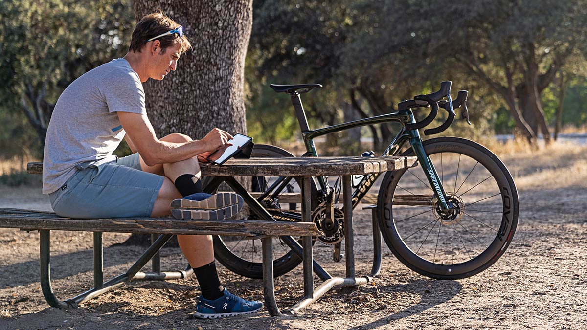Endurance Coach Sitting At Picnic Table Outside Next To Bike Using Trainingpeaks Features On His Laptop