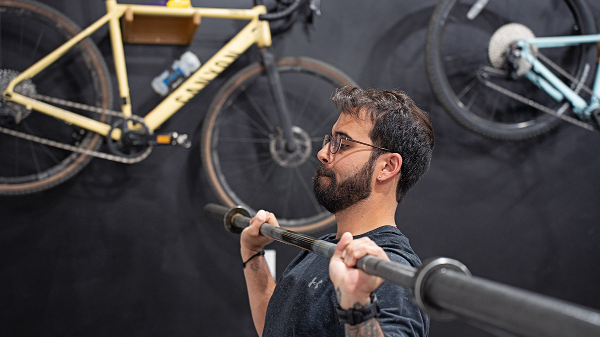 Cycling Athlete In Gym Performing Barbell Press With Bikes On The Wall In Background