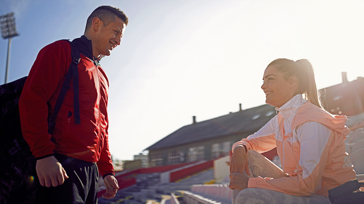 Young Adult Woman Athlete Sitting On Bleachers Smiling And Talking To Male Coach About Menstrual Health