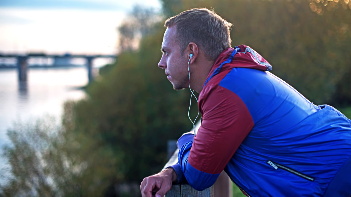 Young Adult Male Athlete Leaning Over Railing Looking Out Into Distance With Headphones In While Taking A Rest Day From Triaining