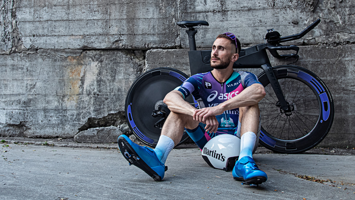 Queer Professional Triathlete Cody Beals Sitting Down In Front Of Bike With Arms Crossed On Legs And Looking Off Into The Distance