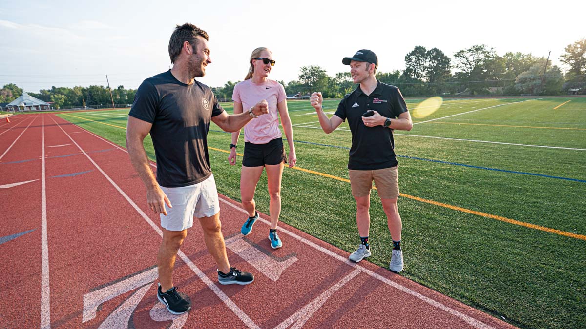 A Male Coach Gives A Fist Bump To A Male Runner As A Female Runner Looks On At A Track.