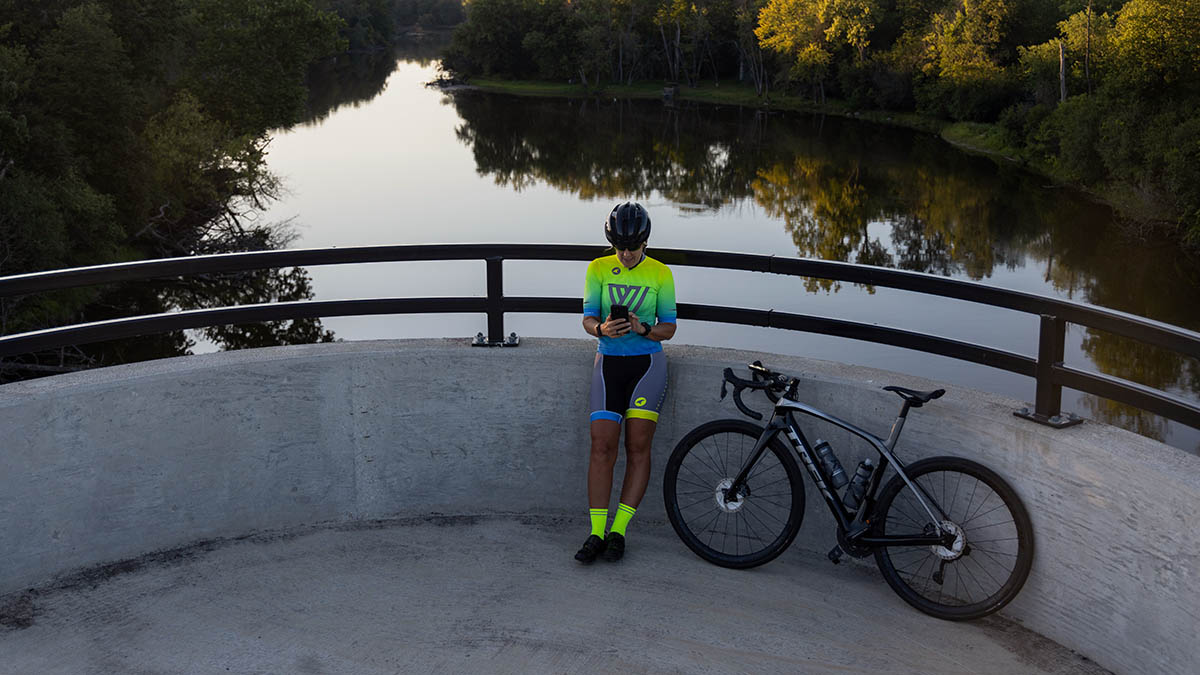 A Female Cyclist At A River Overlook Reviewing Her Data On A Smartphone As The Sun Sets.