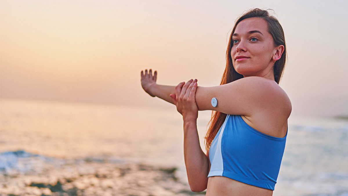 A Woman Stretching Her Shoulder And Arm With A Glucose Monitor On Her Tricep.